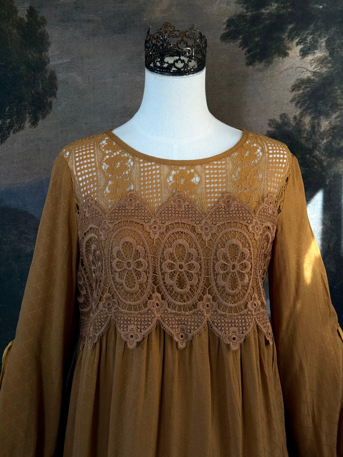 A medieval renaissance inspired tunic dress with cathedral arch lace, billowy slashed bishop sleeves, all in a golden yellow fabric. Pictured on a mannequin in front of a historical painting backdrop. Close up.