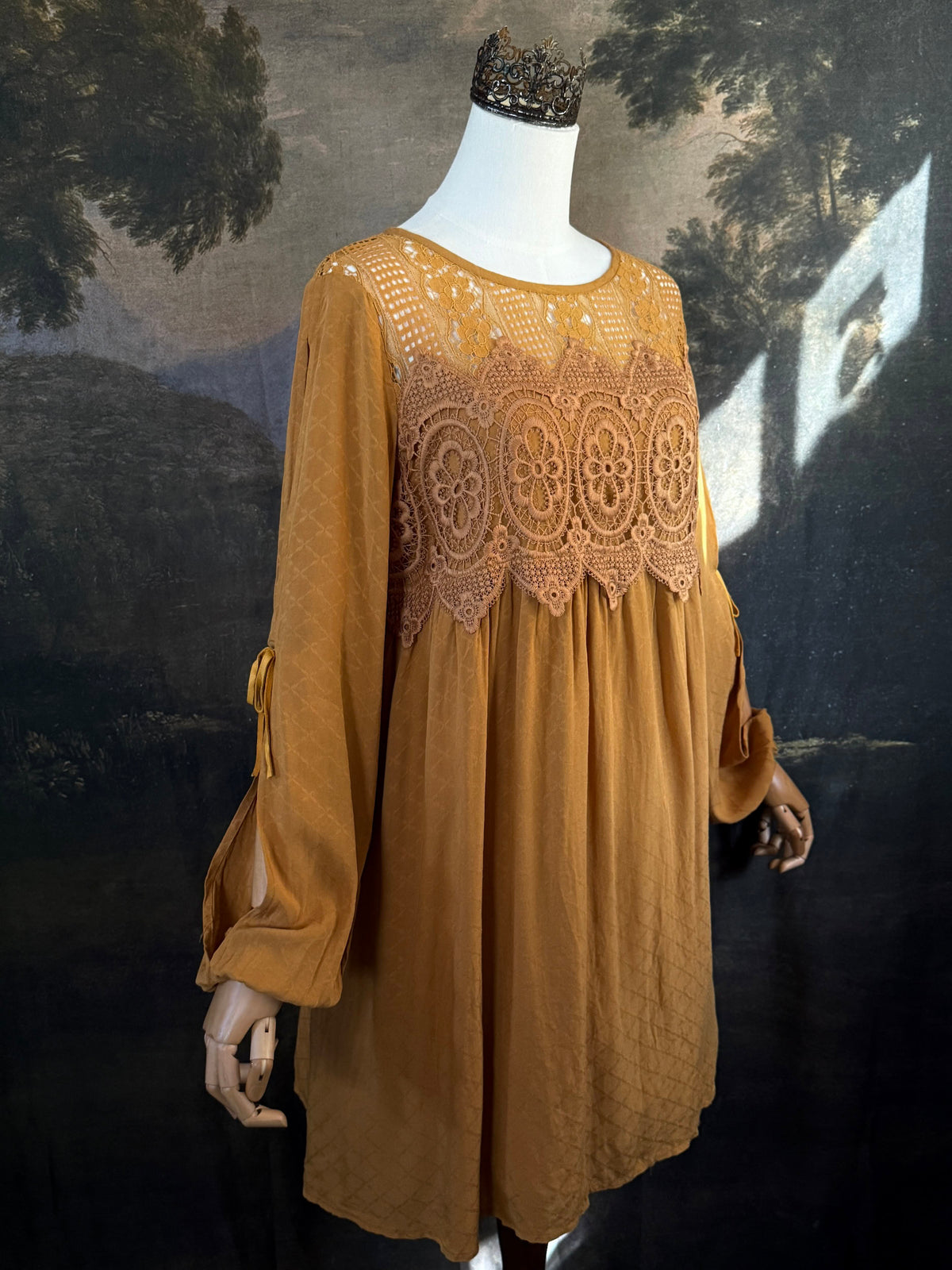A medieval renaissance inspired tunic dress with cathedral arch lace, billowy slashed bishop sleeves, all in a golden yellow fabric. Pictured on a mannequin in front of a historical painting backdrop.