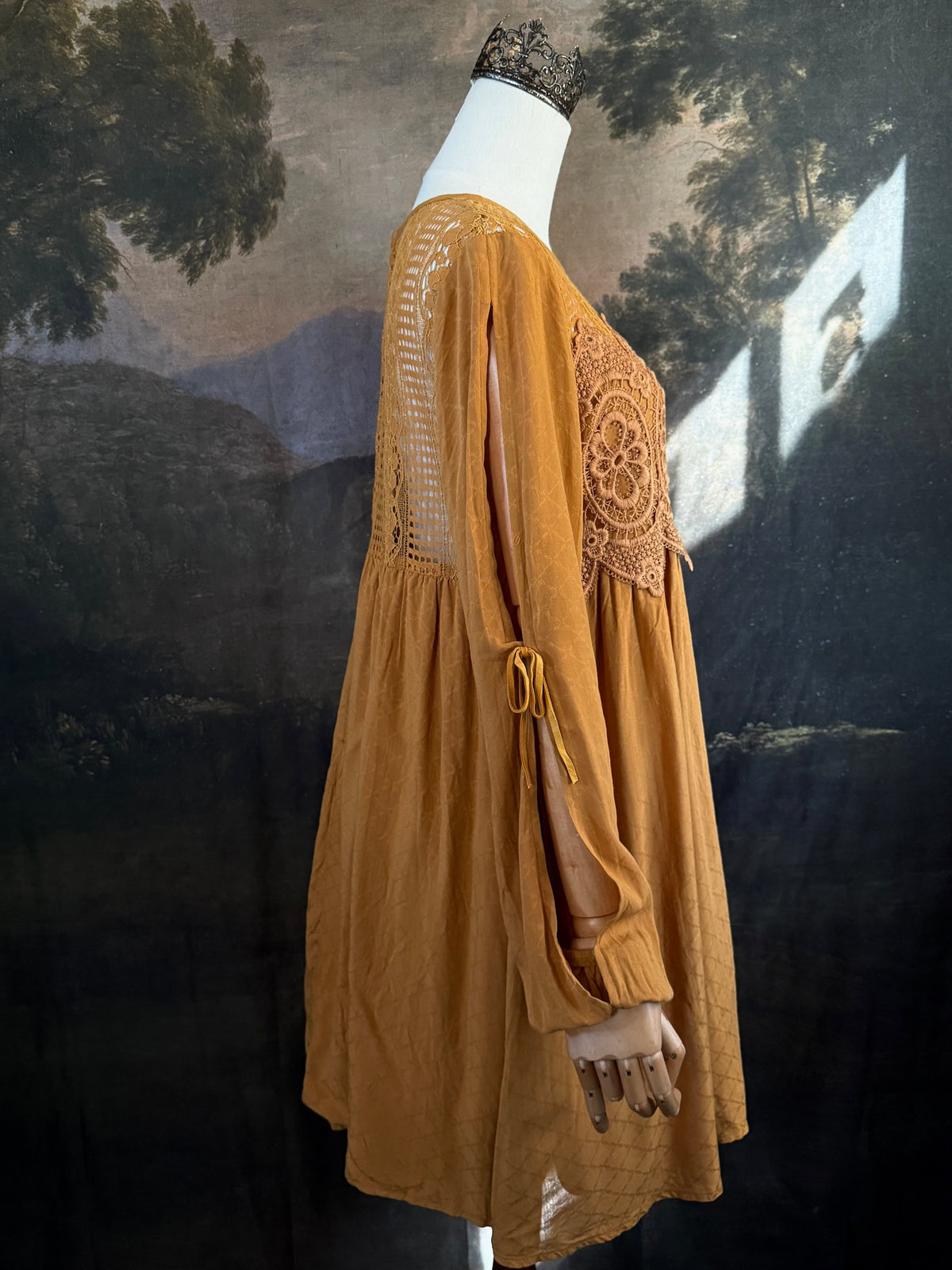 A medieval renaissance inspired tunic dress with cathedral arch lace, billowy slashed bishop sleeves, all in a golden yellow fabric. Pictured on a mannequin in front of a historical painting backdrop.
