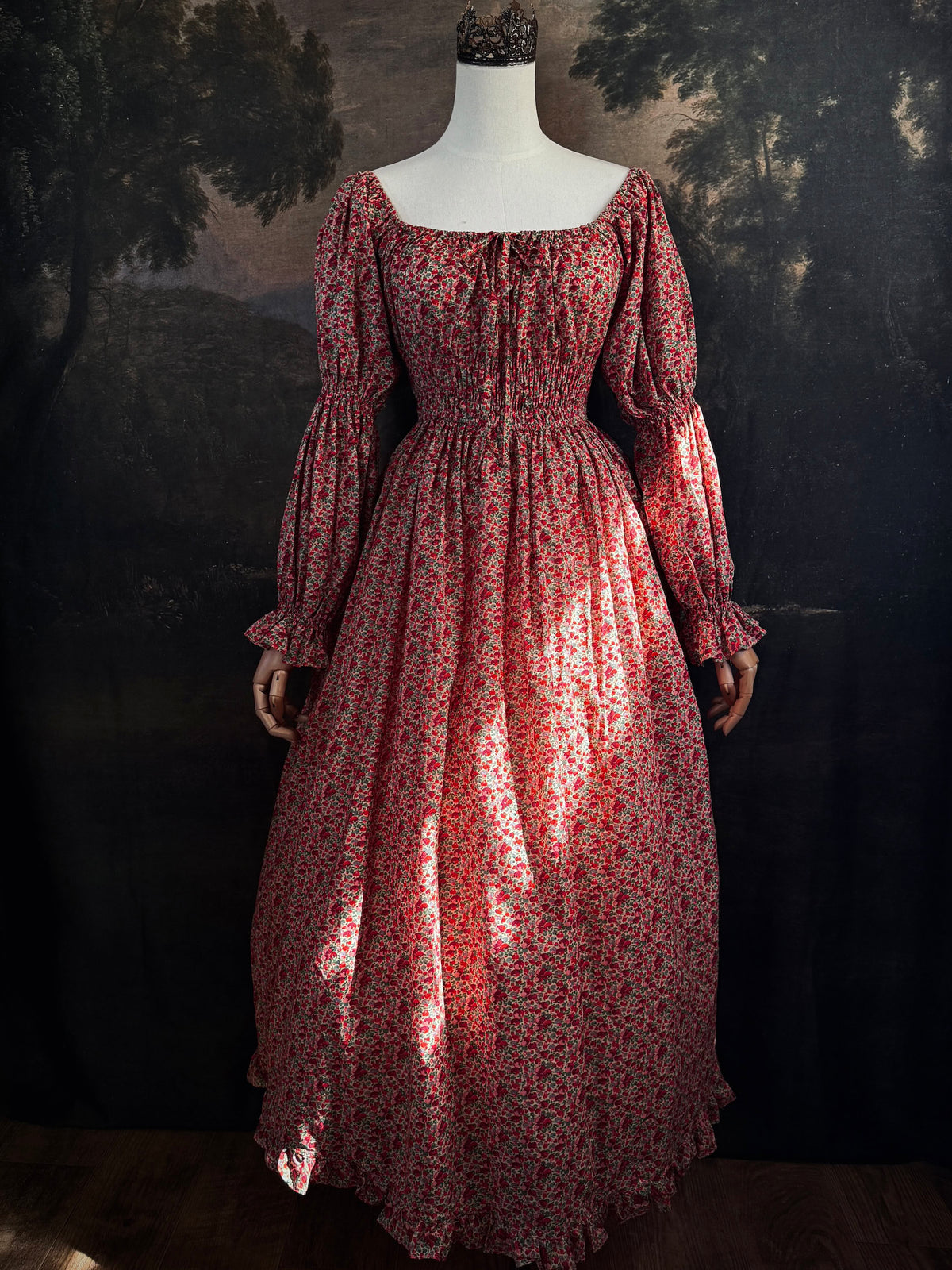 A historically inspired renaissance style, long-sleeved, square-neck drawstring neckline maxi dress in a red apple floral pattern print cotton, with smocked waist and juliet sleeves, displayed on a mannequin against a dark painted  backdrop.