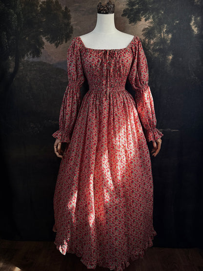 A historically inspired renaissance style, long-sleeved, square-neck drawstring neckline maxi dress in a red apple floral pattern print cotton, with smocked waist and juliet sleeves, displayed on a mannequin against a dark painted  backdrop.