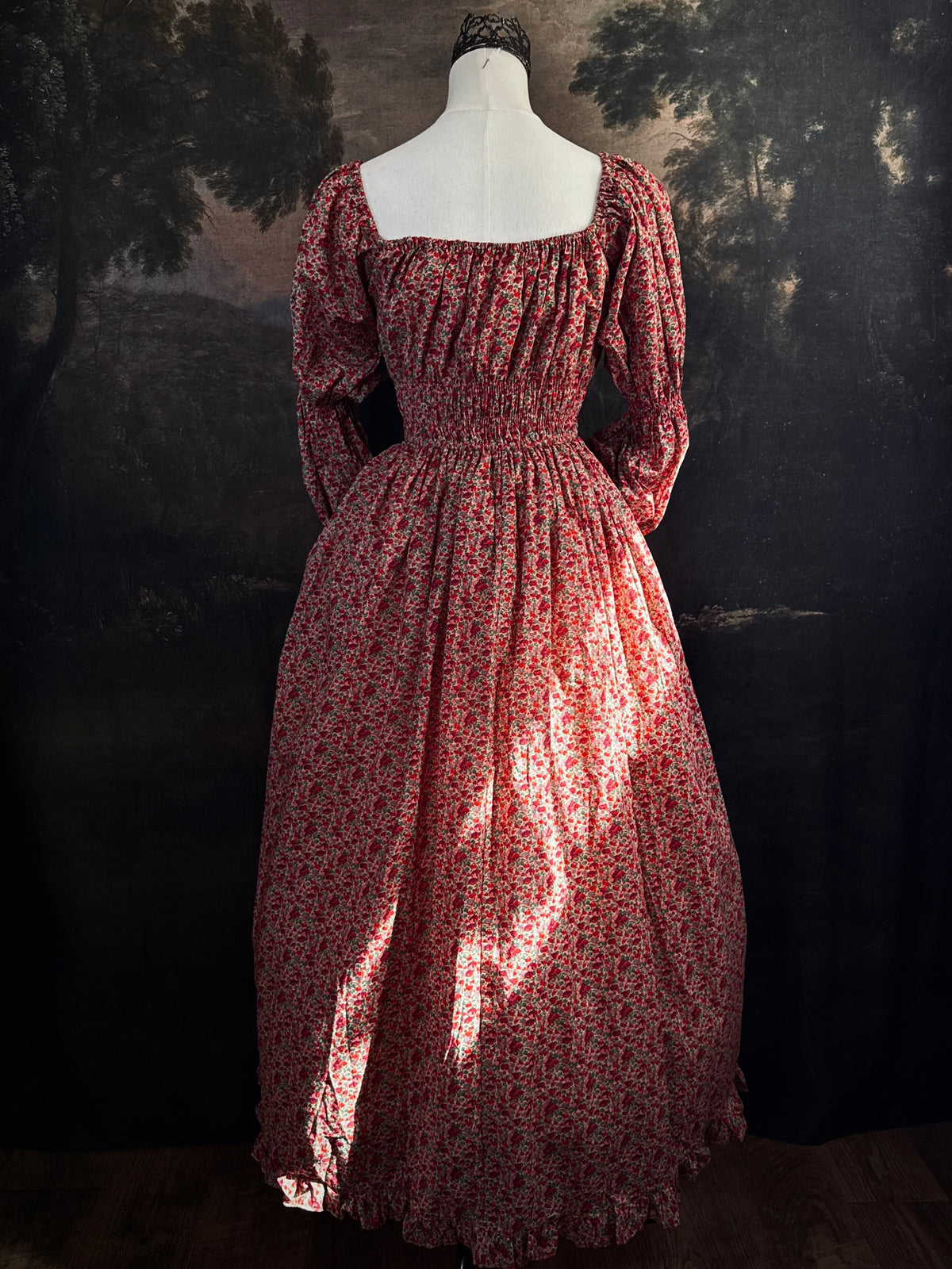 A historically inspired renaissance style, long-sleeved, square-neck drawstring neckline maxi dress in a red apple floral pattern print cotton, with smocked waist and juliet sleeves, displayed on a mannequin against a dark painted  backdrop.