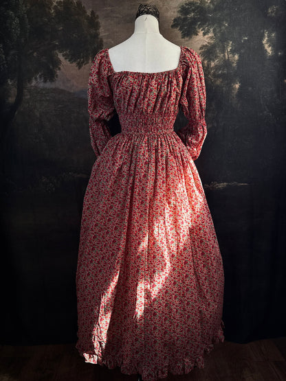A historically inspired renaissance style, long-sleeved, square-neck drawstring neckline maxi dress in a red apple floral pattern print cotton, with smocked waist and juliet sleeves, displayed on a mannequin against a dark painted  backdrop.