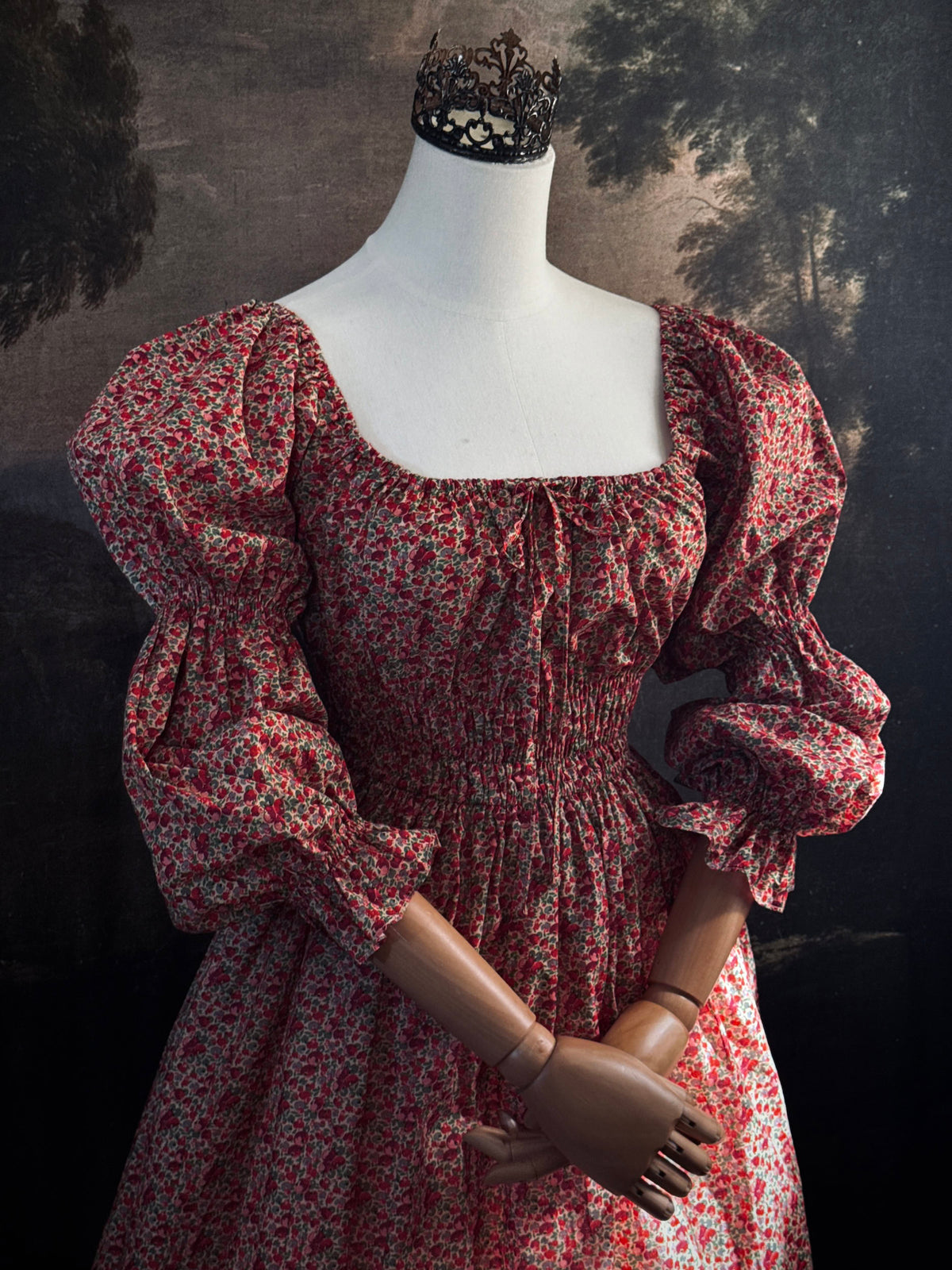 A historically inspired renaissance style, long-sleeved, square-neck drawstring neckline maxi dress in a red apple floral pattern print cotton, with smocked waist and juliet sleeves, displayed on a mannequin against a dark painted  backdrop.