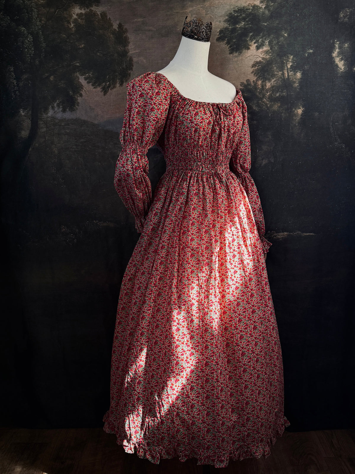 A historically inspired renaissance style, long-sleeved, square-neck drawstring neckline maxi dress in a red apple floral pattern print cotton, with smocked waist and juliet sleeves, displayed on a mannequin against a dark painted  backdrop.
