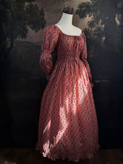 A historically inspired renaissance style, long-sleeved, square-neck drawstring neckline maxi dress in a red apple floral pattern print cotton, with smocked waist and juliet sleeves, displayed on a mannequin against a dark painted  backdrop.