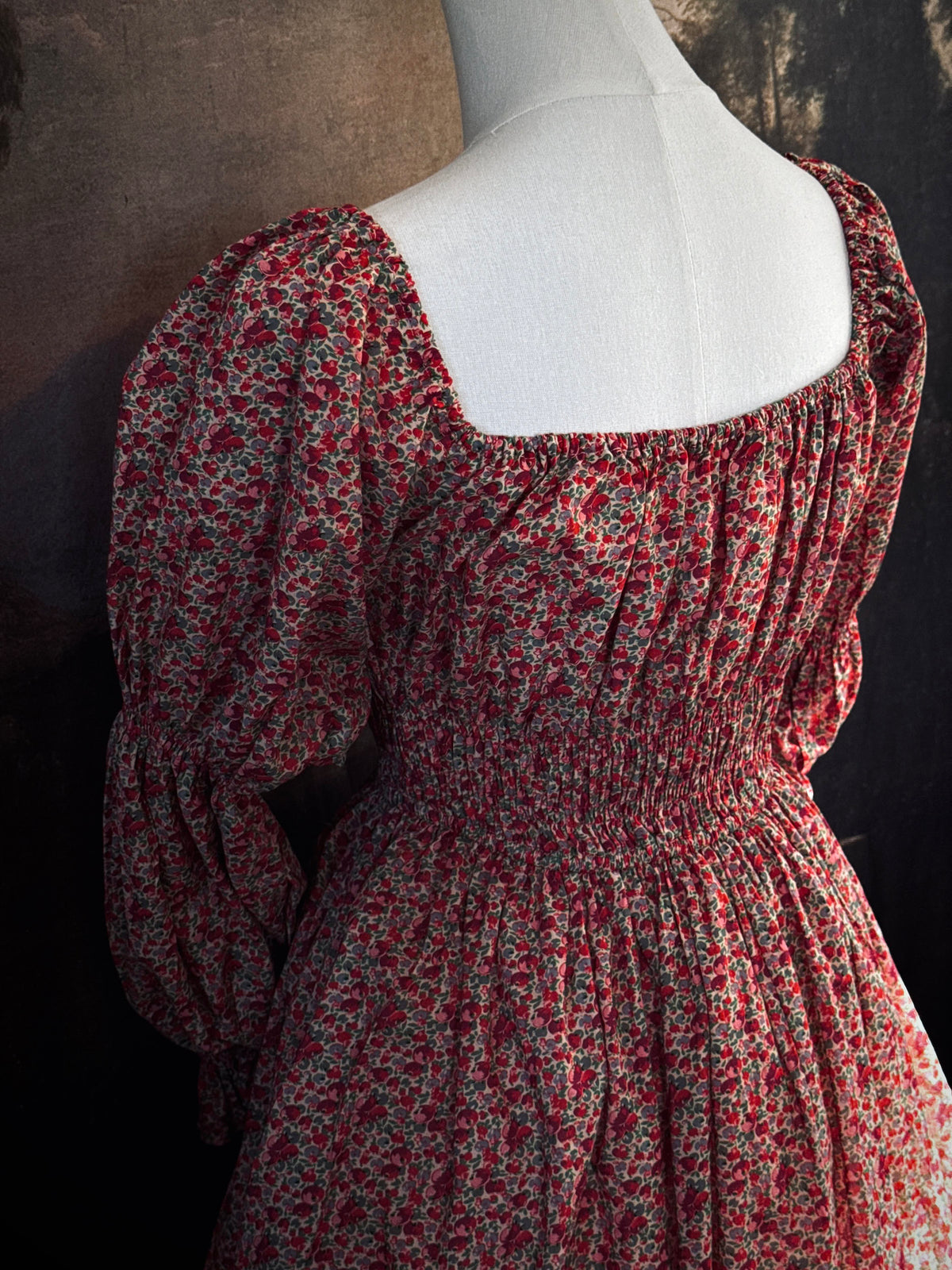A historically inspired renaissance style, long-sleeved, square-neck drawstring neckline maxi dress in a red apple floral pattern print cotton, with smocked waist and juliet sleeves, displayed on a mannequin against a dark painted  backdrop.
