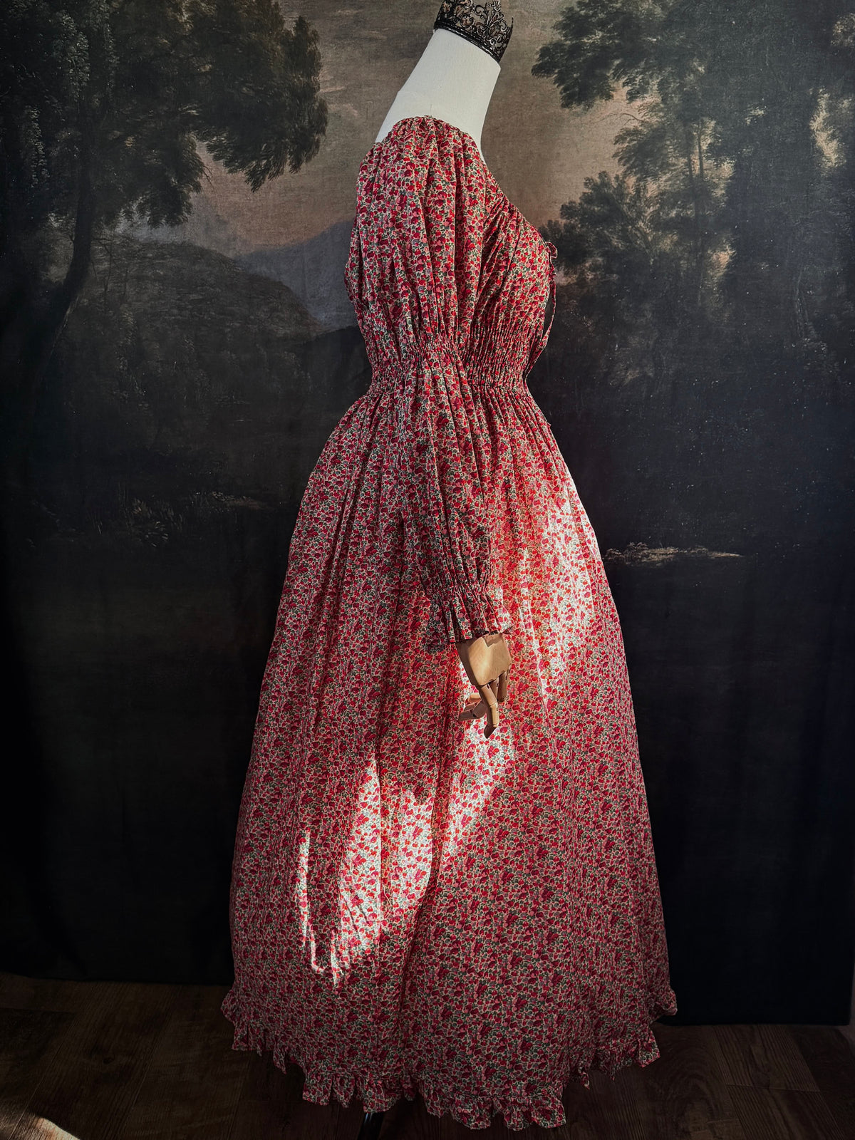 A historically inspired renaissance style, long-sleeved, square-neck drawstring neckline maxi dress in a red apple floral pattern print cotton, with smocked waist and juliet sleeves, displayed on a mannequin against a dark painted  backdrop.