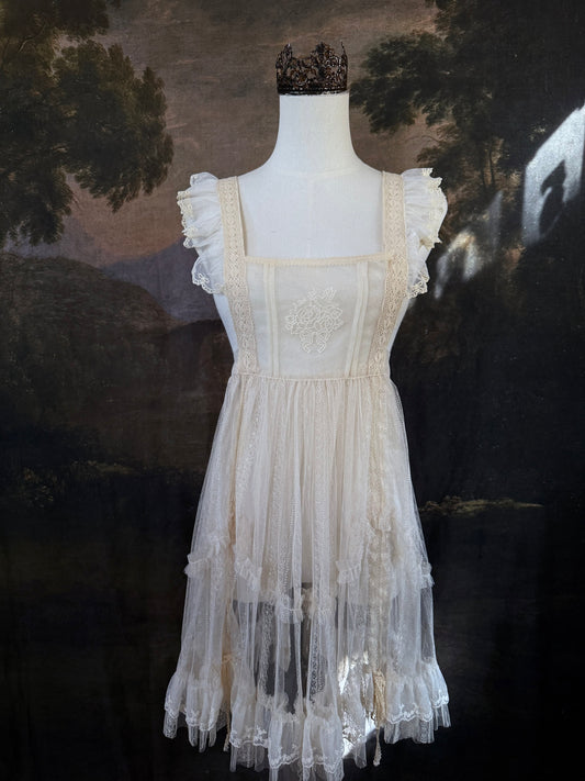 An antique-style cream lace apron over-dress displayed on a mannequin, featuring ruffled details, embroidery, a square neckline, tiered tulle skirt, set against a dark classical landscape painting background.