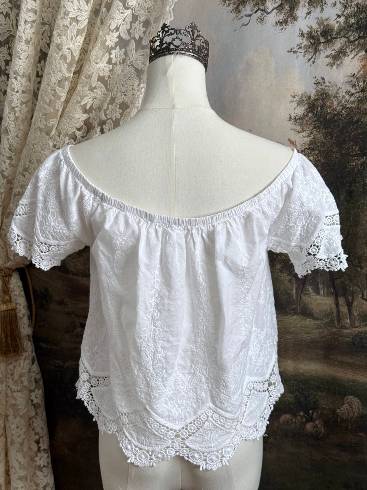 A historically inspired white embroidered peasant chemise blouse with short sleeves on a mannequin in front of an art history painting backdrop of a field