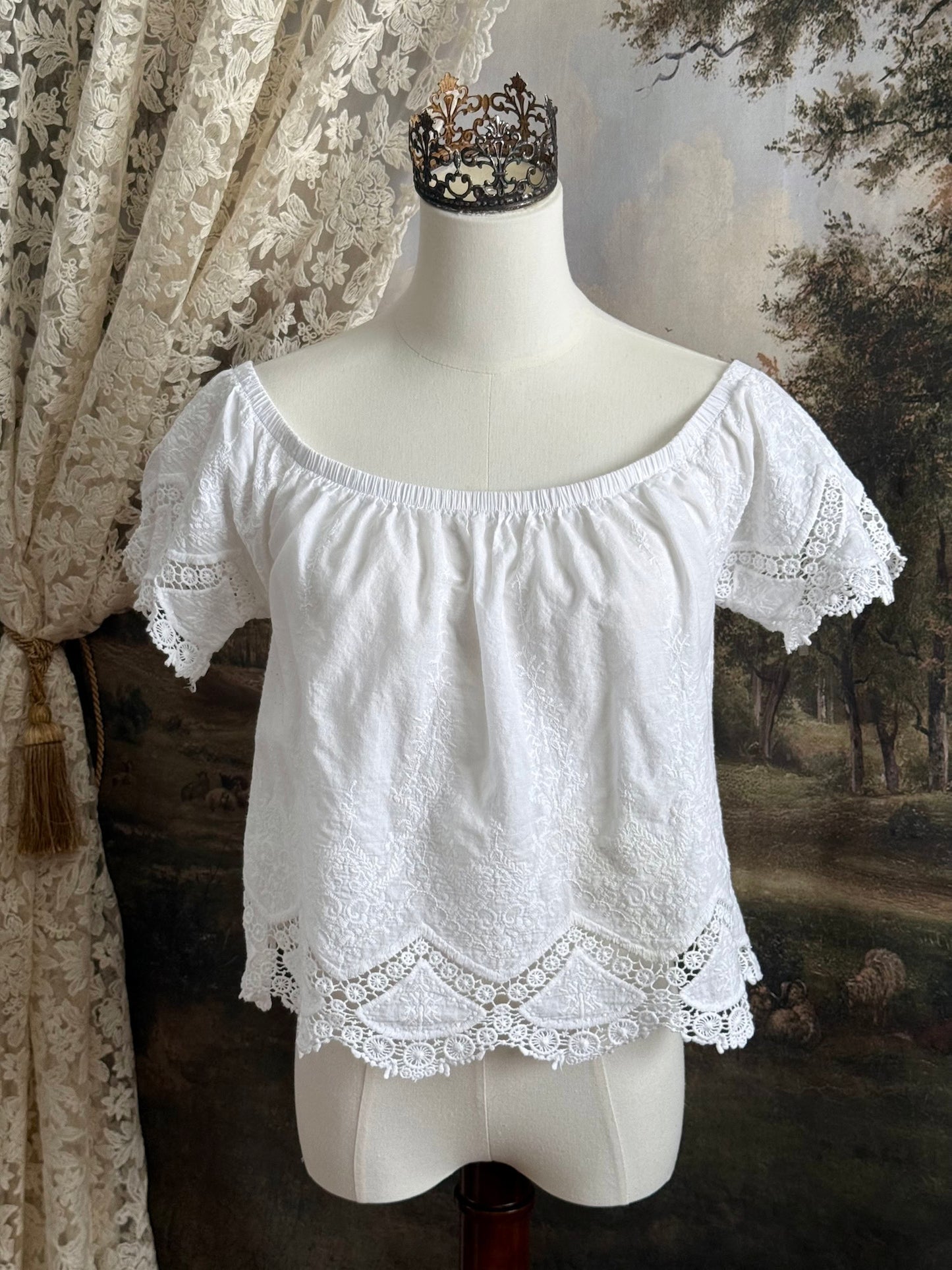 A historically inspired white embroidered peasant chemise blouse with short sleeves on a mannequin in front of an art history painting backdrop of a field