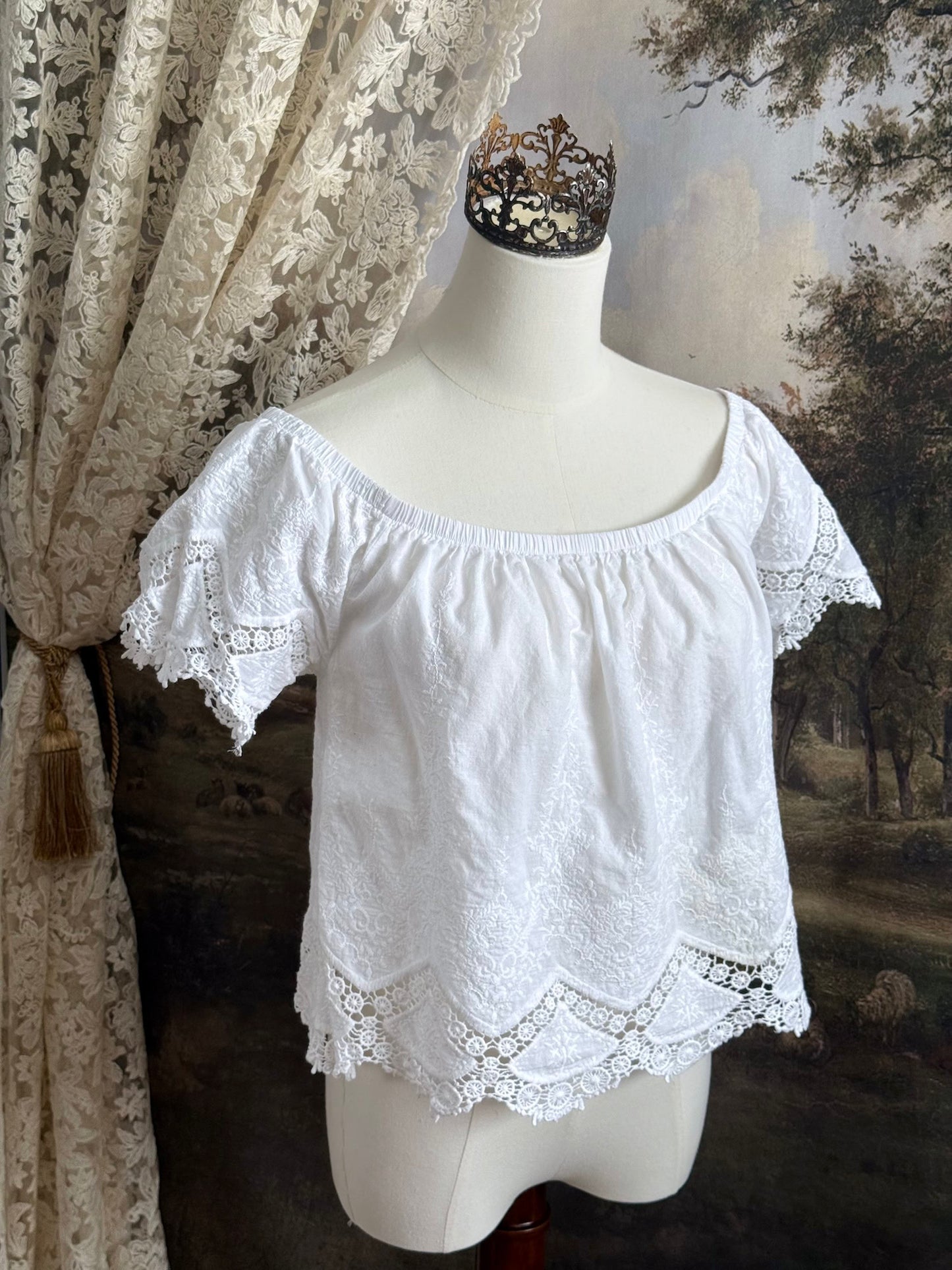 A historically inspired white embroidered peasant chemise blouse with short sleeves on a mannequin in front of an art history painting backdrop of a field
