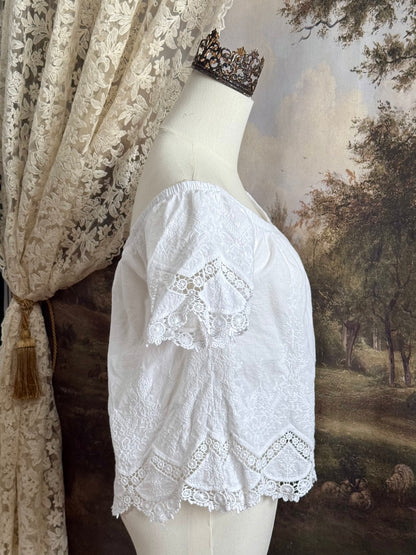 A historically inspired white embroidered peasant chemise blouse with short sleeves on a mannequin in front of an art history painting backdrop of a field