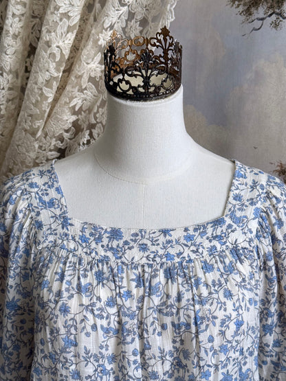A white and blue historically inspired floral vine print blouse with square neckline and bishop sleeves, pictured on a mannequin in front of an art history backdrop