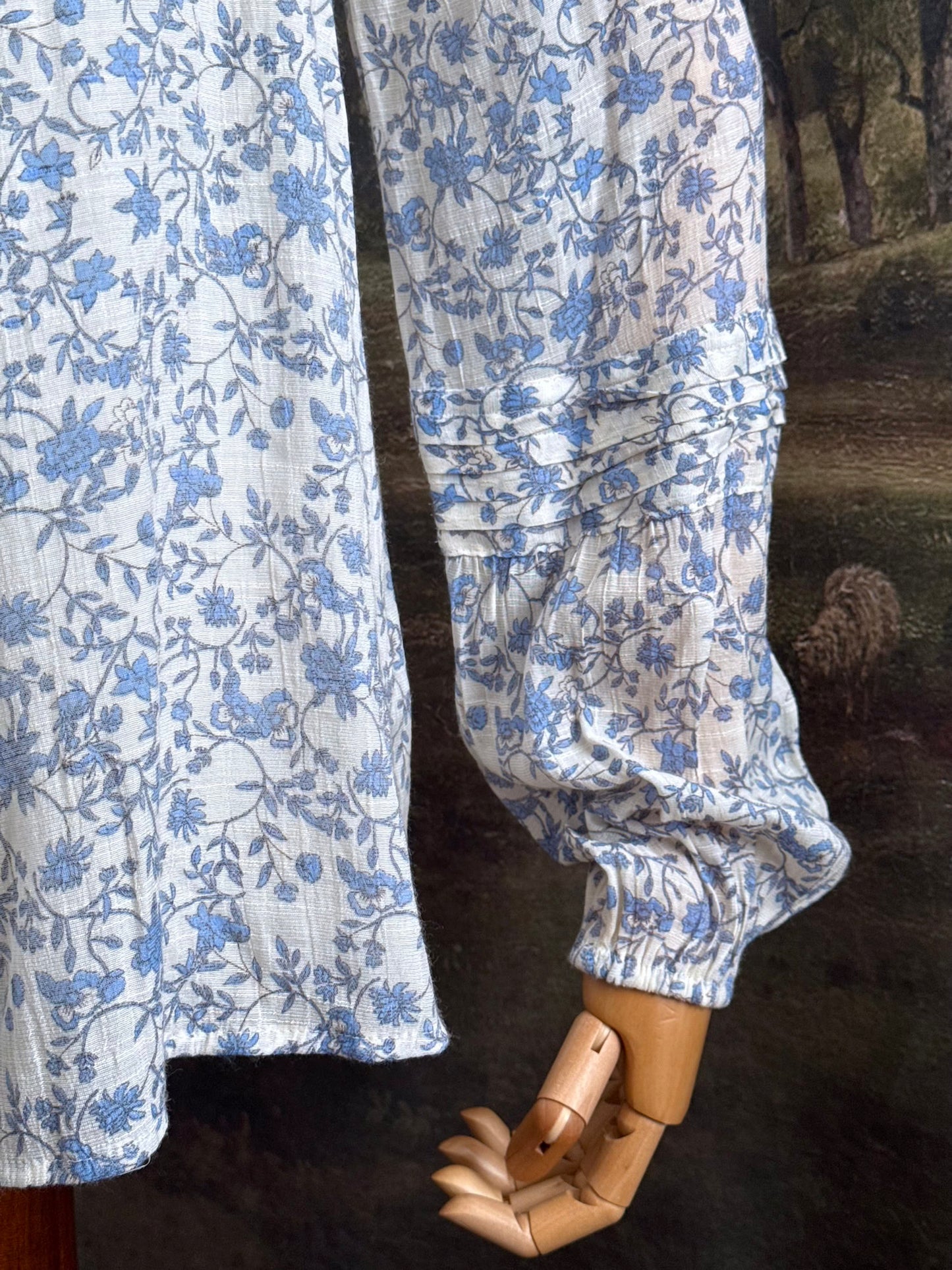 A white and blue historically inspired floral vine print blouse with square neckline and bishop sleeves, pictured on a mannequin in front of an art history backdrop