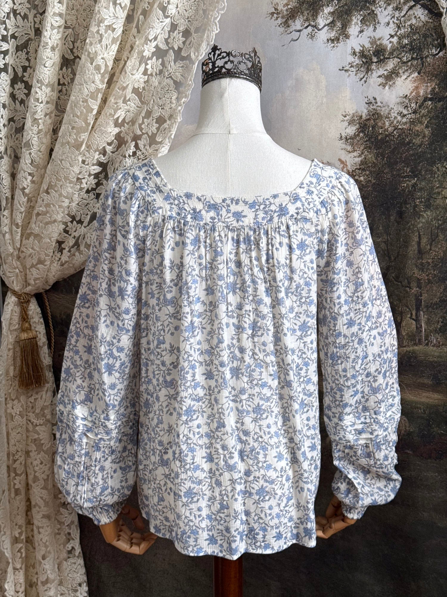 A white and blue historically inspired floral vine print blouse with square neckline and bishop sleeves, pictured on a mannequin in front of an art history backdrop
