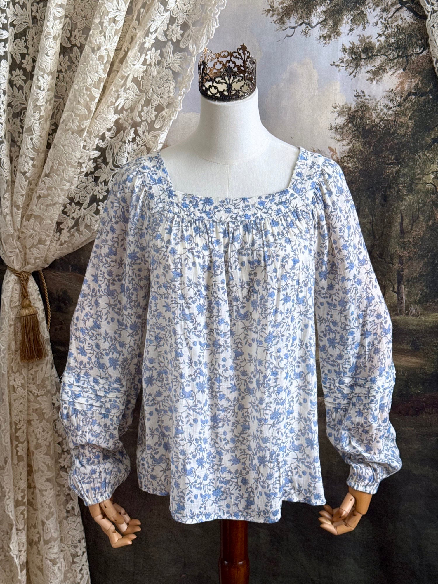 A white and blue historically inspired floral vine print blouse with square neckline and bishop sleeves, pictured on a mannequin in front of an art history backdrop