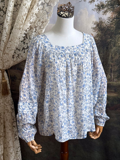 A white and blue historically inspired floral vine print blouse with square neckline and bishop sleeves, pictured on a mannequin in front of an art history backdrop