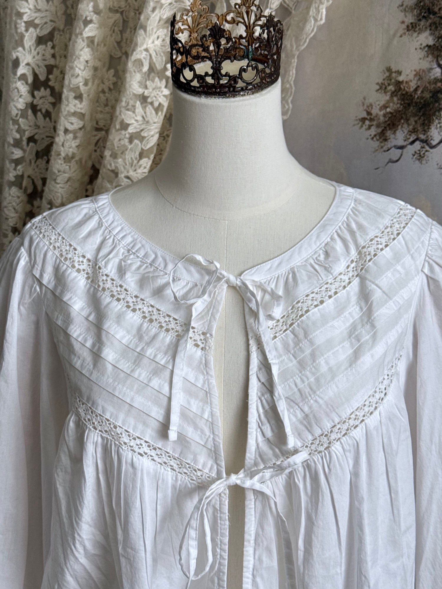 Historically inspired white cotton blouse with bishop sleeves, pintuck pleated details, lace trim, and a tie-front sillhouette. Pictured on a mannequin in front of an art history backdrop.