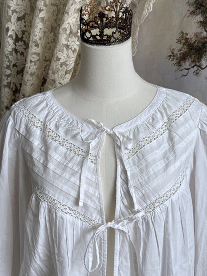 Historically inspired white cotton blouse with bishop sleeves, pintuck pleated details, lace trim, and a tie-front sillhouette. Pictured on a mannequin in front of an art history backdrop.
