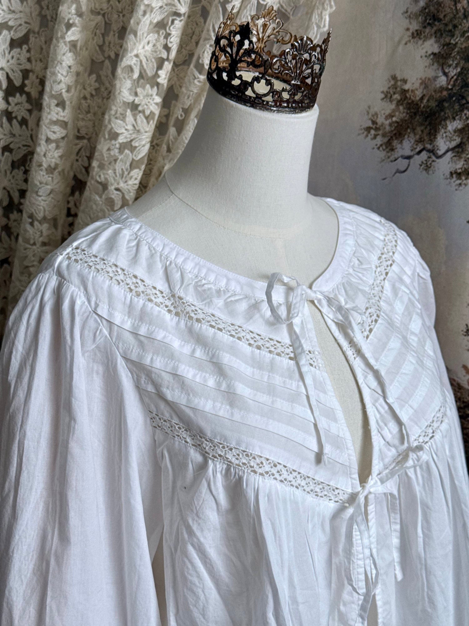 Historically inspired white cotton blouse with bishop sleeves, pintuck pleated details, lace trim, and a tie-front sillhouette. Pictured on a mannequin in front of an art history backdrop.
