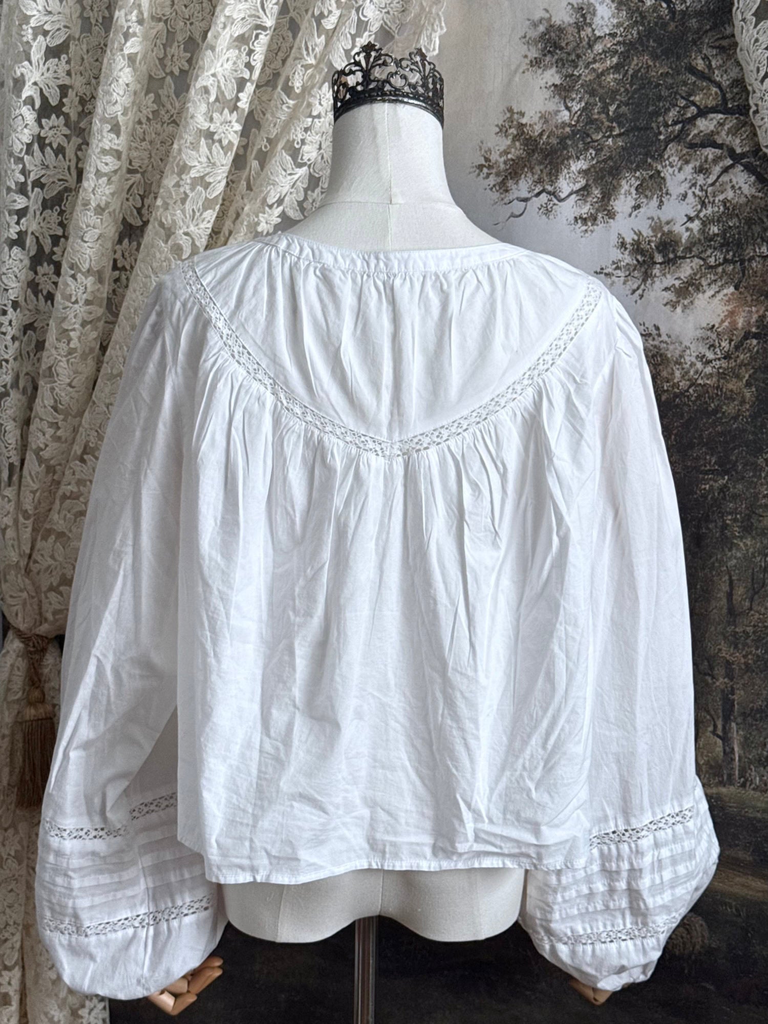 Historically inspired white cotton blouse with bishop sleeves, pintuck pleated details, lace trim, and a tie-front sillhouette. Pictured on a mannequin in front of an art history backdrop.