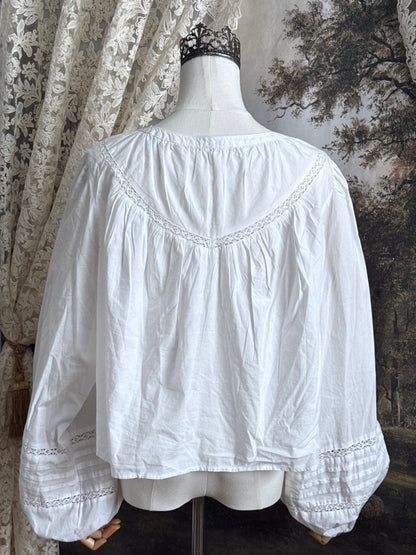 Historically inspired white cotton blouse with bishop sleeves, pintuck pleated details, lace trim, and a tie-front sillhouette. Pictured on a mannequin in front of an art history backdrop.