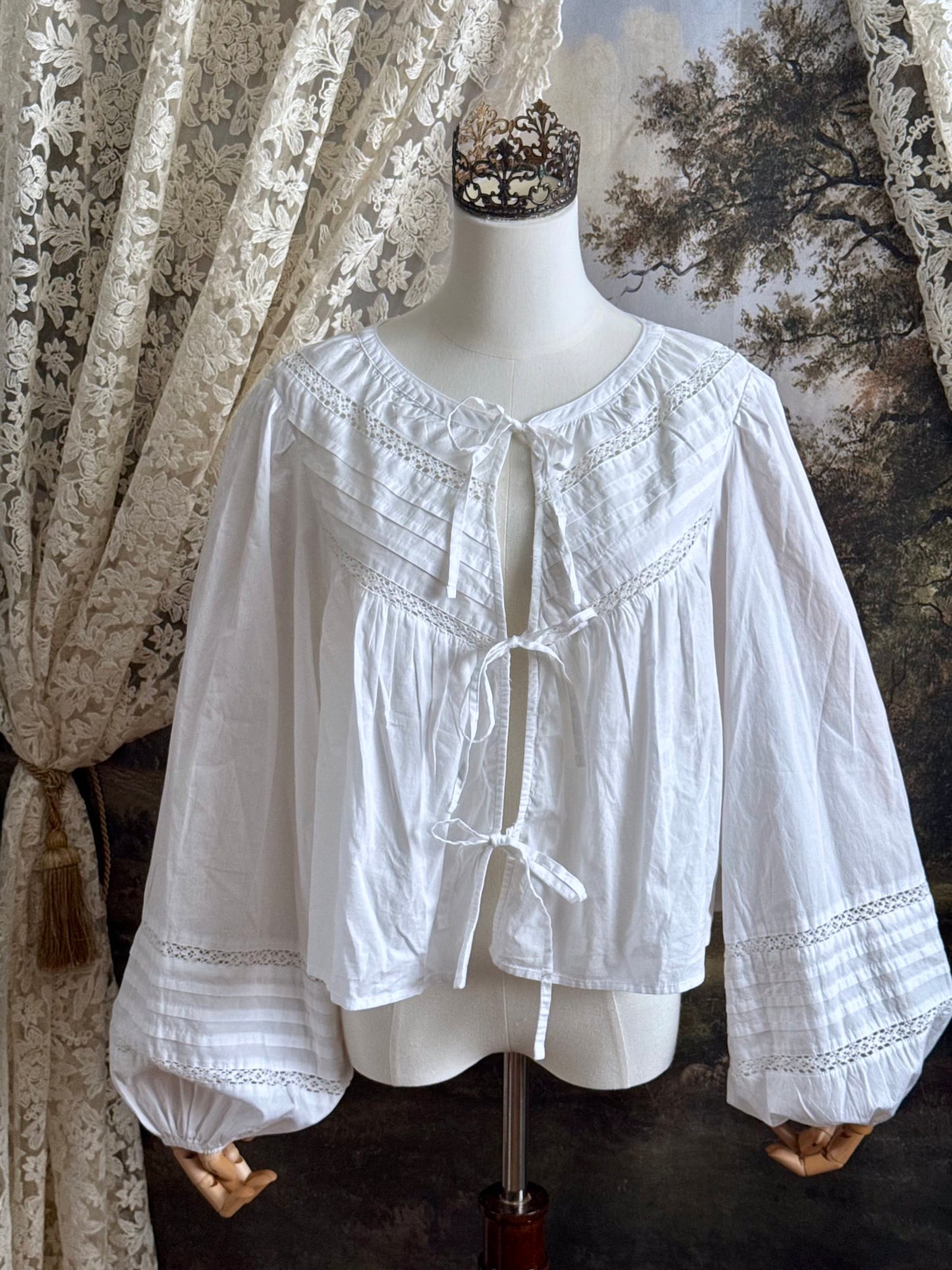 Historically inspired white cotton blouse with bishop sleeves, pintuck pleated details, lace trim, and a tie-front sillhouette. Pictured on a mannequin in front of an art history backdrop.