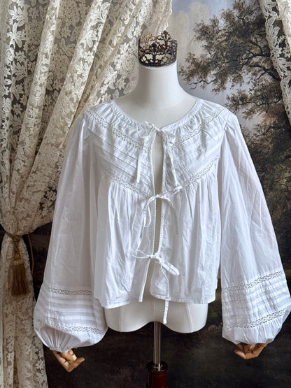 Historically inspired white cotton blouse with bishop sleeves, pintuck pleated details, lace trim, and a tie-front sillhouette. Pictured on a mannequin in front of an art history backdrop.