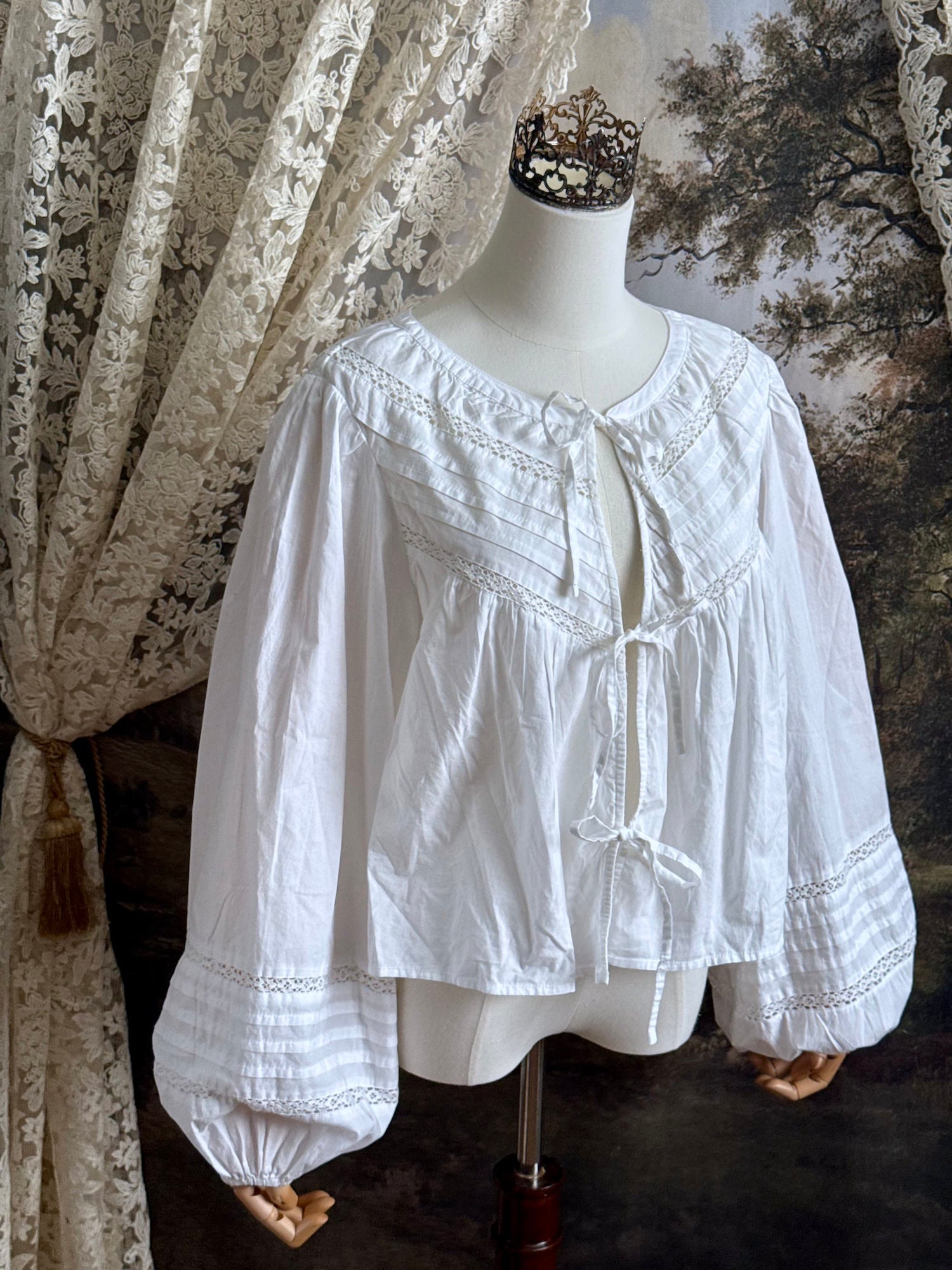 Historically inspired white cotton blouse with bishop sleeves, pintuck pleated details, lace trim, and a tie-front sillhouette. Pictured on a mannequin in front of an art history backdrop.