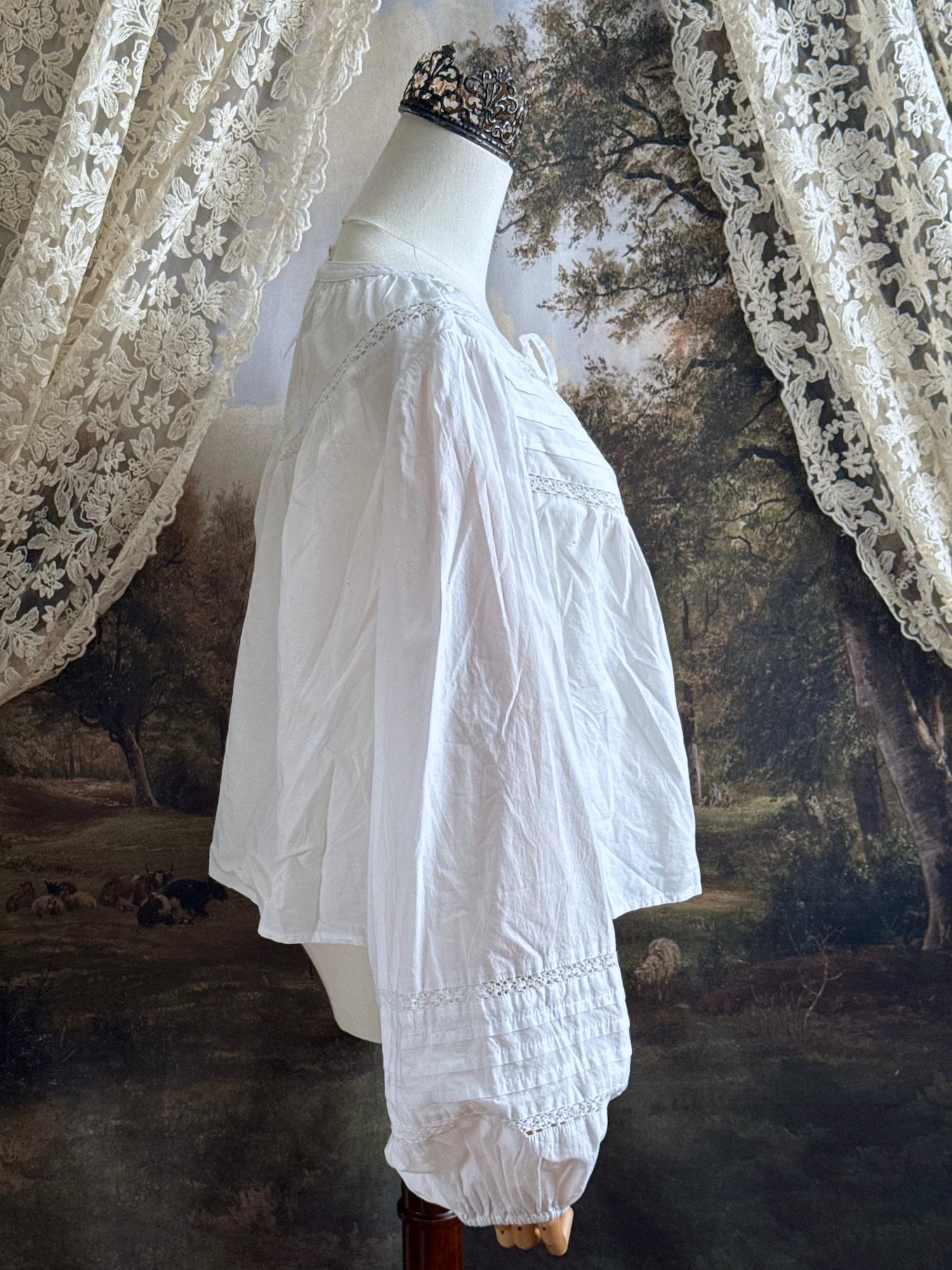 Historically inspired white cotton blouse with bishop sleeves, pintuck pleated details, lace trim, and a tie-front sillhouette. Pictured on a mannequin in front of an art history backdrop.