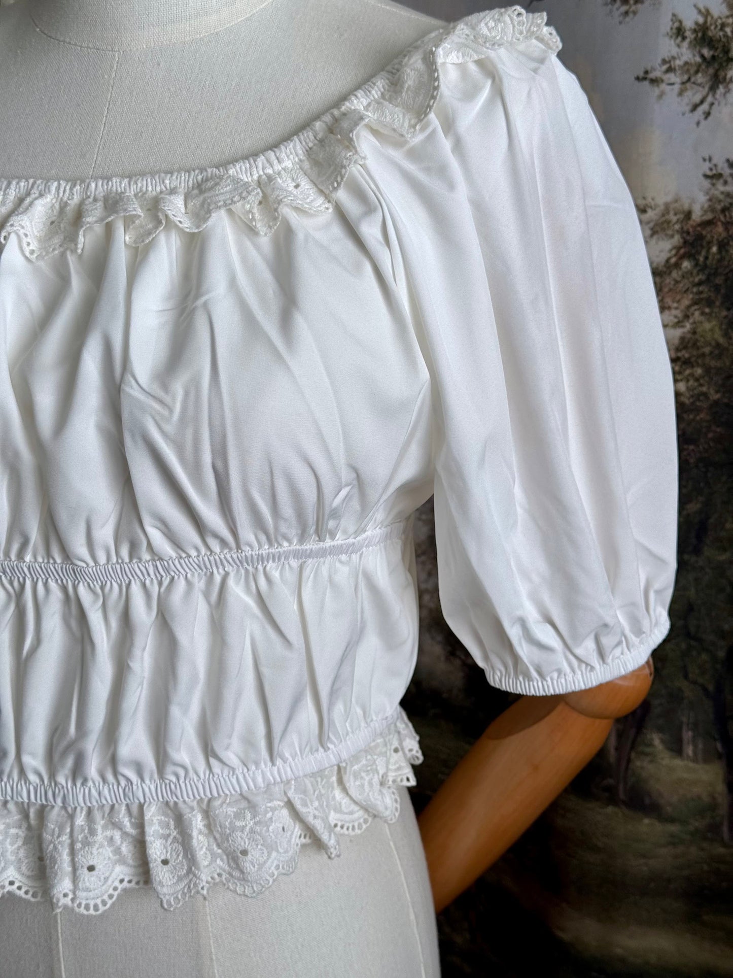 Historical fashion inspired vintage style white shirred top chemise with puff sleeves and floral lace trim on a mannequin in front of an art history backdrop and lace curtain. Detail of sleeve.