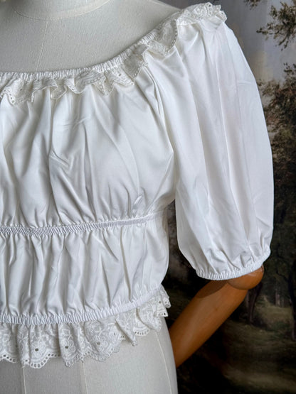 Historical fashion inspired vintage style white shirred top chemise with puff sleeves and floral lace trim on a mannequin in front of an art history backdrop and lace curtain. Detail of sleeve.