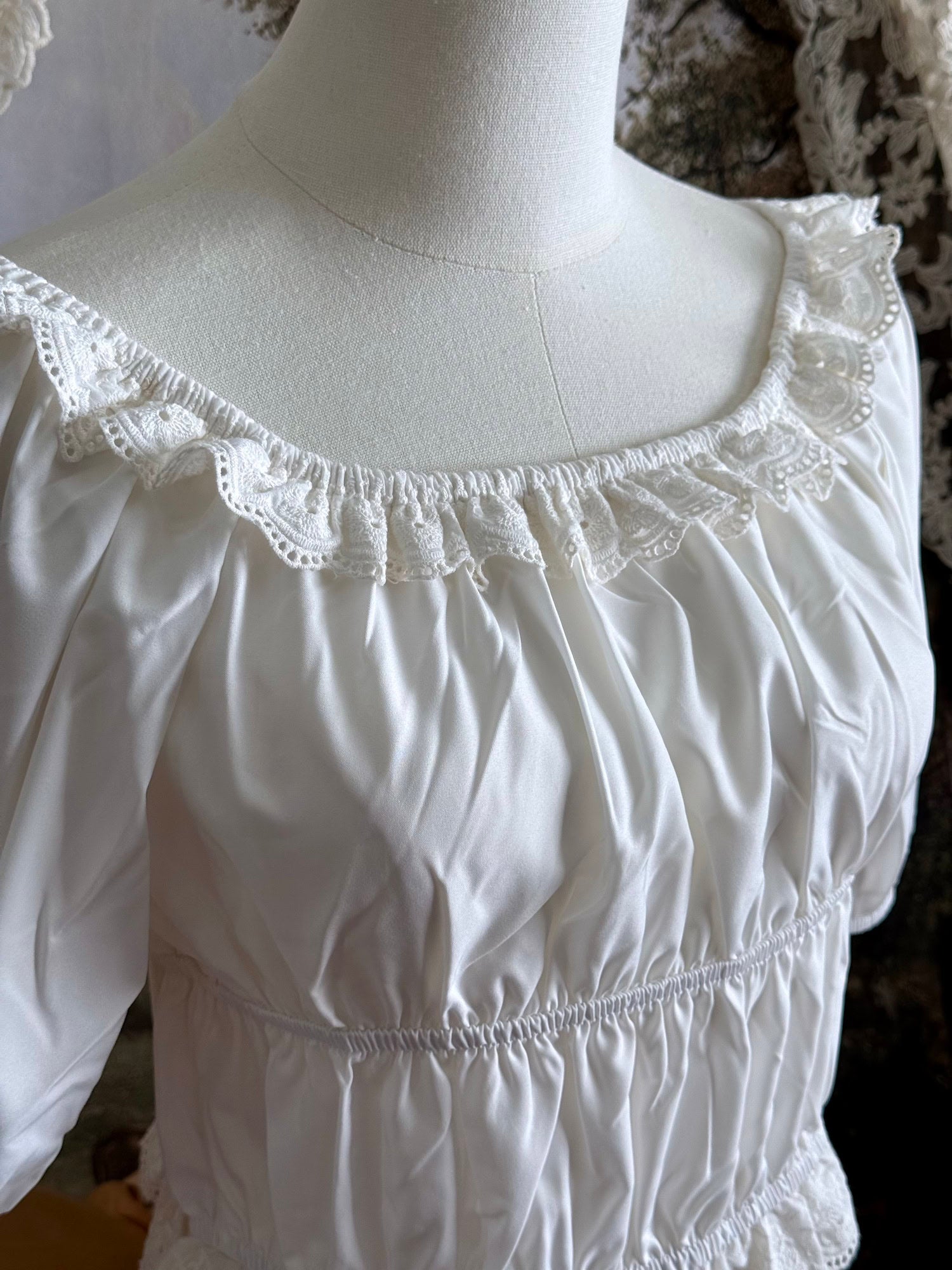 Historical fashion inspired vintage style white shirred top chemise with puff sleeves and floral lace trim on a mannequin in front of an art history backdrop and lace curtain. Detail shot.