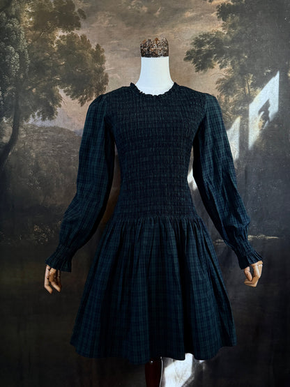 A dark navy blue and green tartan print smocked long-sleeve drop waist dress with a pleated skirt, displayed on a mannequin against a classical landscape painting backdrop.
