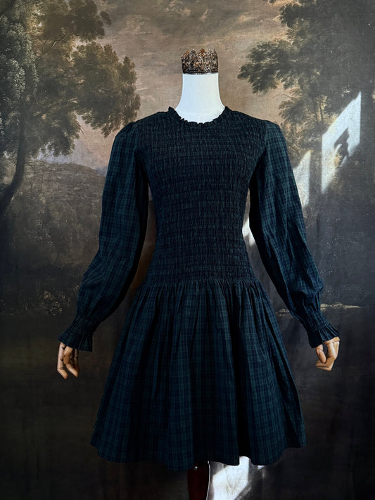 A dark navy blue and green tartan print smocked long-sleeve drop waist dress with a pleated skirt, displayed on a mannequin against a classical landscape painting backdrop.