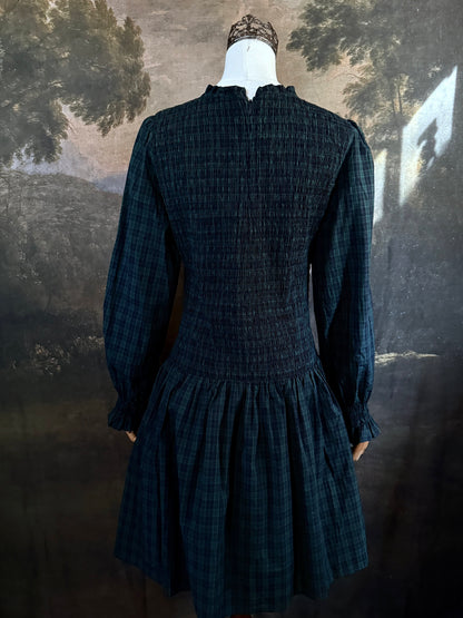 A dark navy blue and green tartan print smocked long-sleeve drop waist dress with a pleated skirt, displayed on a mannequin against a classical landscape painting backdrop.