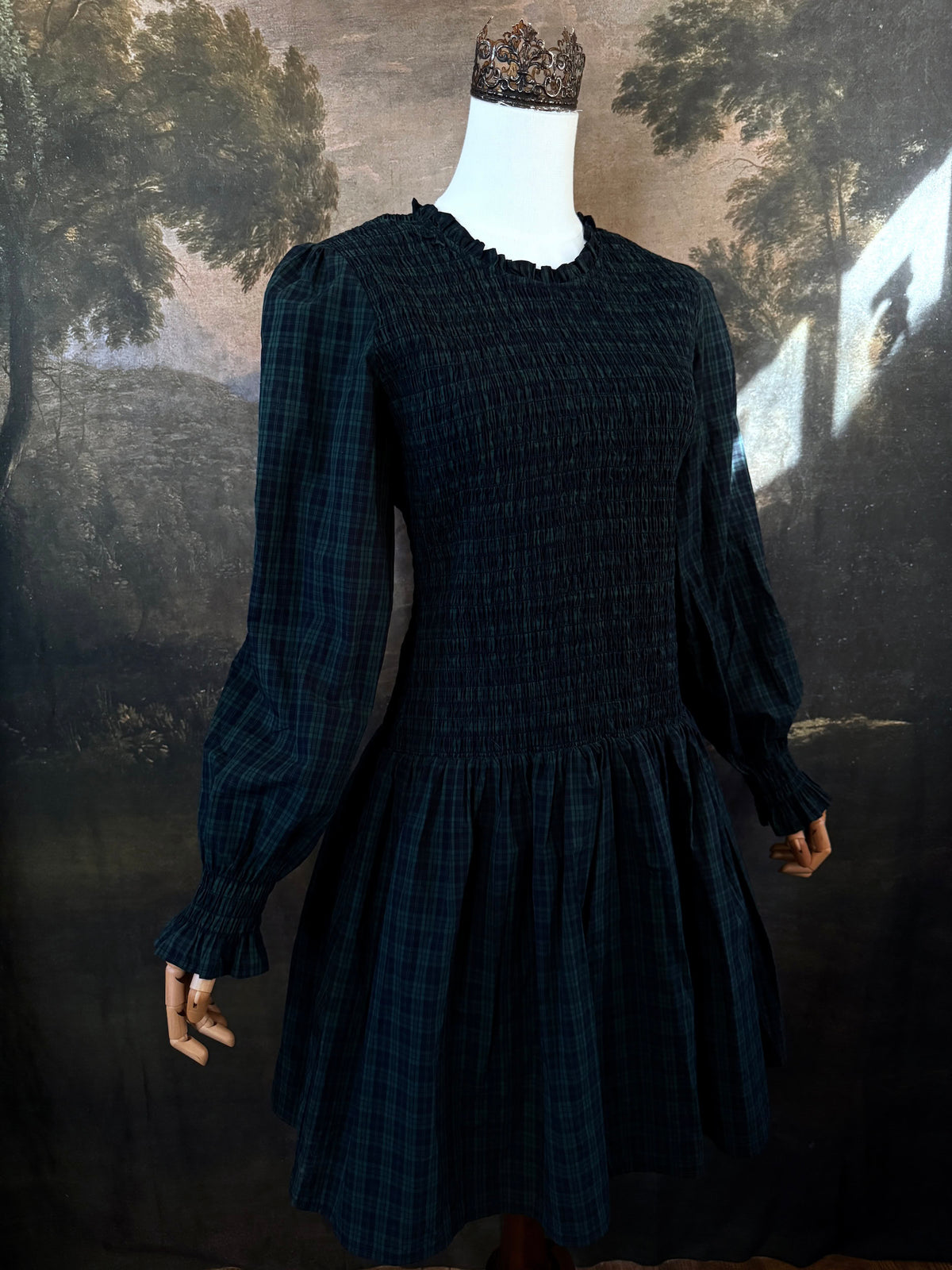 A dark navy blue and green tartan print smocked long-sleeve drop waist dress with a pleated skirt, displayed on a mannequin against a classical landscape painting backdrop.