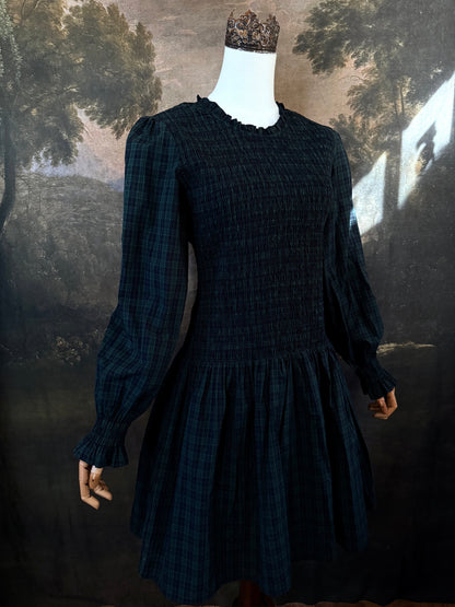 A dark navy blue and green tartan print smocked long-sleeve drop waist dress with a pleated skirt, displayed on a mannequin against a classical landscape painting backdrop.