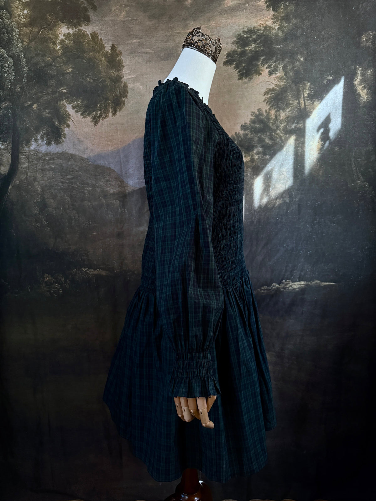 A dark navy blue and green tartan print smocked long-sleeve drop waist dress with a pleated skirt, displayed on a mannequin against a classical landscape painting backdrop.