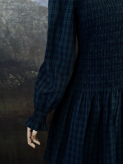 A dark navy blue and green tartan print smocked long-sleeve drop waist dress with a pleated skirt, displayed on a mannequin against a classical landscape painting backdrop.