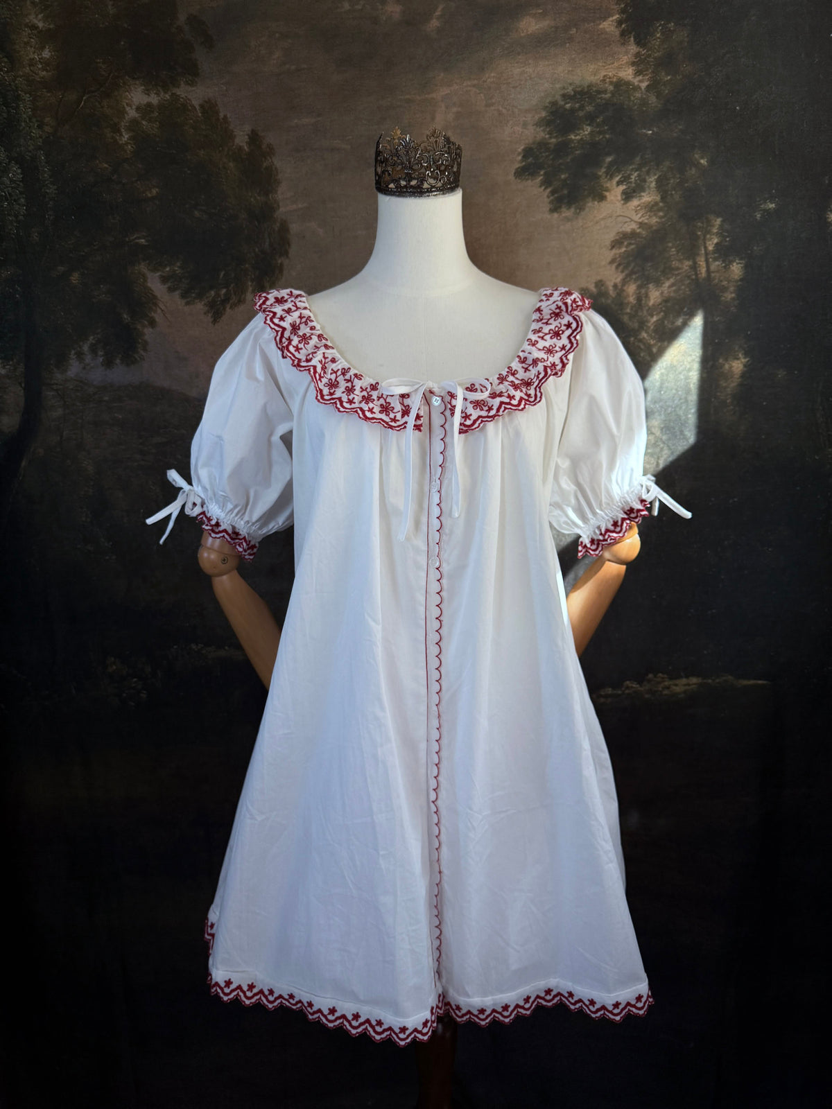 A white historical-style mini chemise dress babydoll style nightgown with dark red embroidered floral trim, displayed on a mannequin against a dark forest painting backdrop.