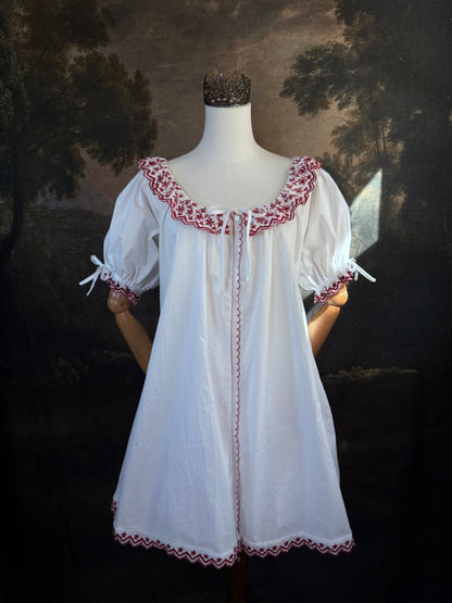 A white historical-style mini chemise dress babydoll style nightgown with dark red embroidered floral trim, displayed on a mannequin against a dark forest painting backdrop.