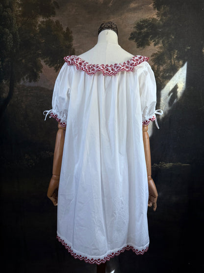 A white historical-style mini chemise dress babydoll style nightgown with dark red embroidered floral trim, displayed on a mannequin against a dark forest painting backdrop.