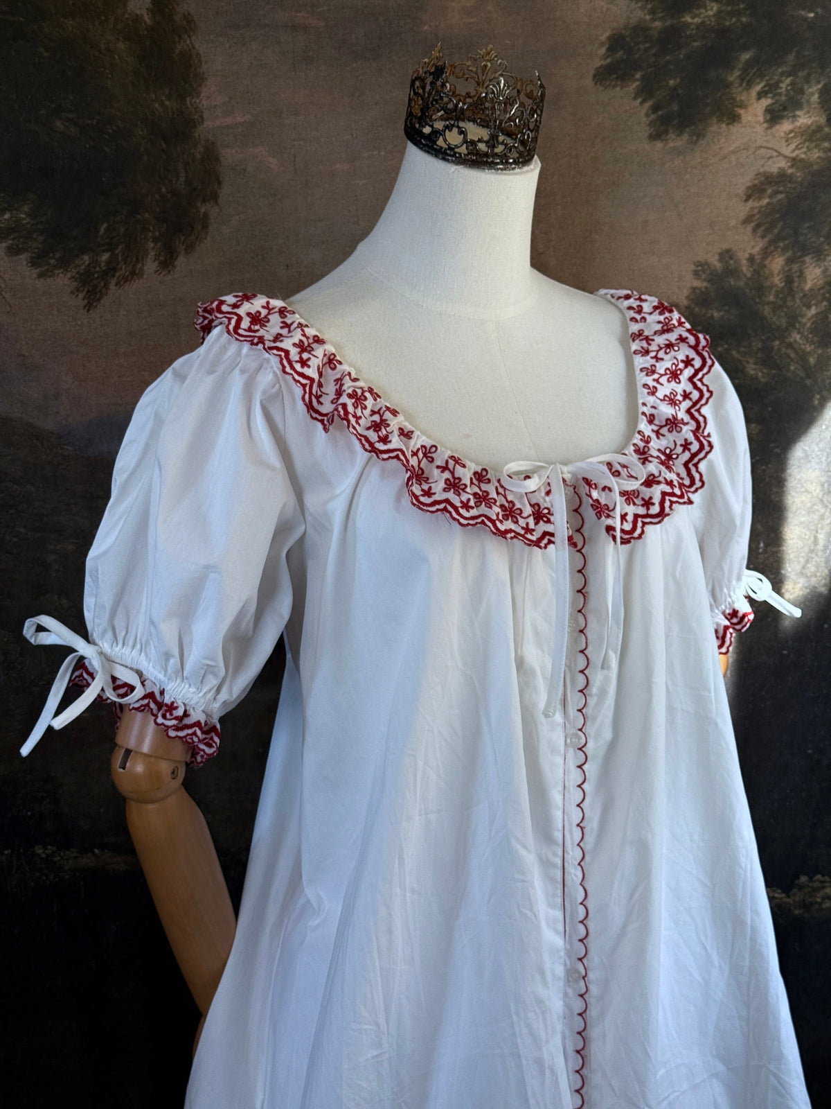 A white historical-style mini chemise dress babydoll style nightgown with dark red embroidered floral trim, displayed on a mannequin against a dark forest painting backdrop.