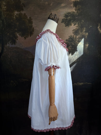A white historical-style mini chemise dress babydoll style nightgown with dark red embroidered floral trim, displayed on a mannequin against a dark forest painting backdrop.