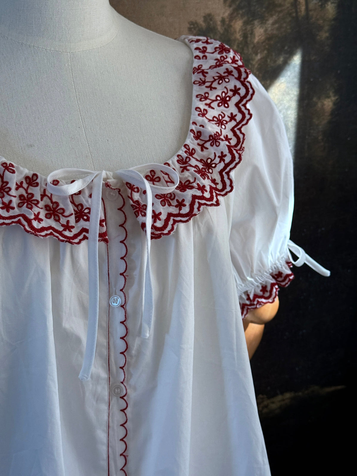 A white historical-style mini chemise dress babydoll style nightgown with dark red embroidered floral trim, displayed on a mannequin against a dark forest painting backdrop.