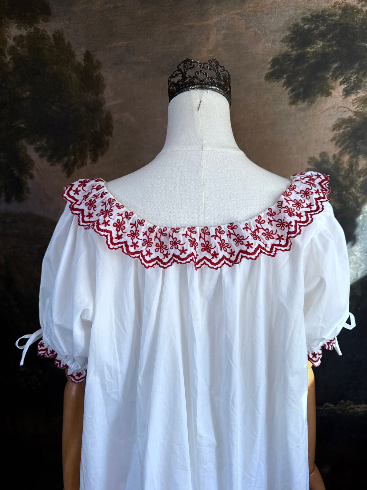 A white historical-style mini chemise dress babydoll style nightgown with dark red embroidered floral trim, displayed on a mannequin against a dark forest painting backdrop.