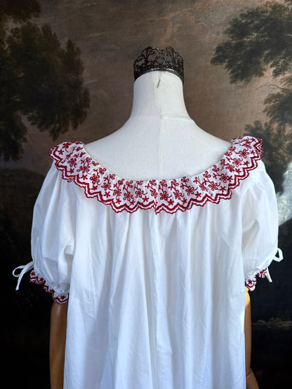 A white historical-style mini chemise dress babydoll style nightgown with dark red embroidered floral trim, displayed on a mannequin against a dark forest painting backdrop.