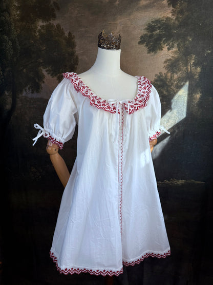 A white historical-style mini chemise dress babydoll style nightgown with dark red embroidered floral trim, displayed on a mannequin against a dark forest painting backdrop.
